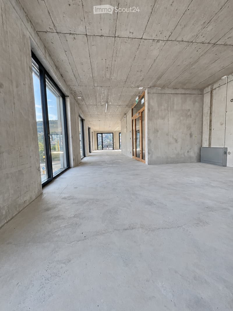 Empty concrete corridor with large windows on one side, leading to another area with more windows and a view of the outdoors.