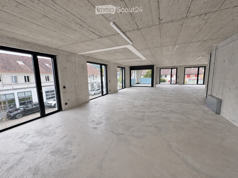 An empty, modern corridor with concrete walls and floors, large glass windows, and recessed lighting.