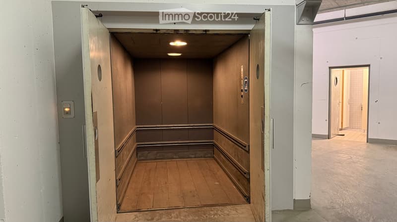 Empty hallway with wooden flooring, white walls, and an open elevator shaft with wooden paneling and ceiling lights.
