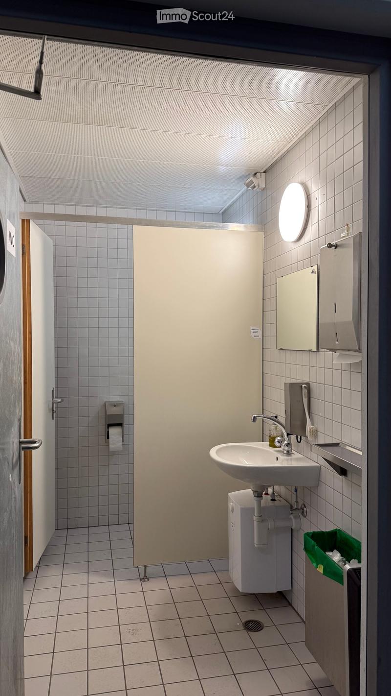 Modern bathroom with white tiled walls and floor, a white sink with a modern faucet, a round mirror, a toilet paper dispenser, and a soap dispenser. A closed stall door is visible in the background.