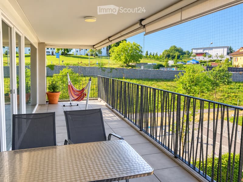 Balcony with a metal table, two chairs, a hammock, and a potted plant, overlooking a green landscape with buildings and trees.