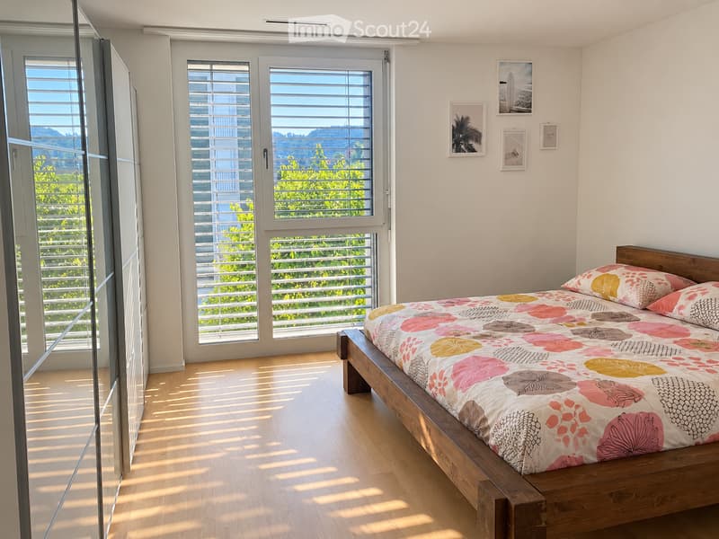 Bright, modern bedroom with wooden flooring, large windows, and a bed with a colorful floral duvet.