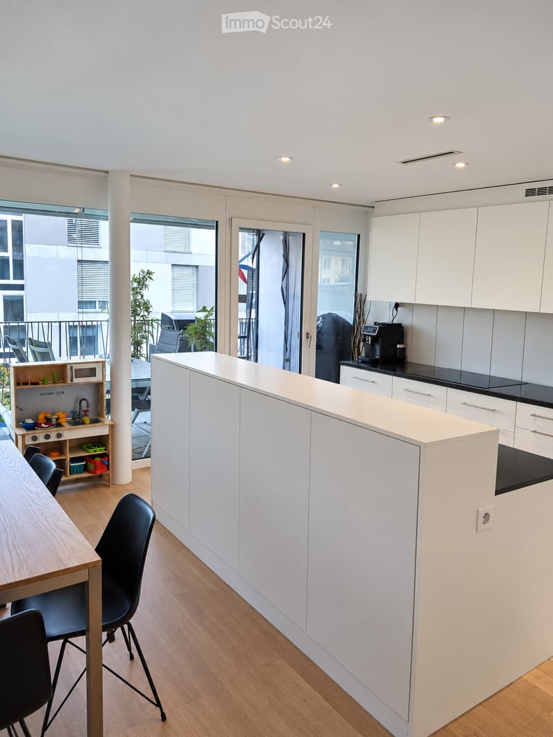 Modern kitchen with white cabinets, wooden floor, island, and a small play kitchen area visible in the background.
