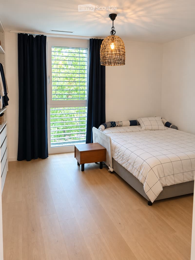 Modern bedroom with light wooden flooring, a white bed with a grid-patterned duvet, a small wooden nightstand, and a wicker pendant light hanging from the ceiling. Large window with dark curtains offers a view of greenery outside.