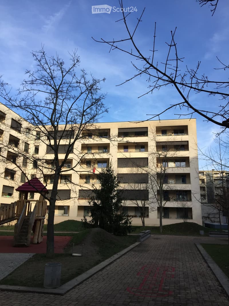 Modern multi-story residential building with balconies and large windows, surrounded by trees and a small playground area.