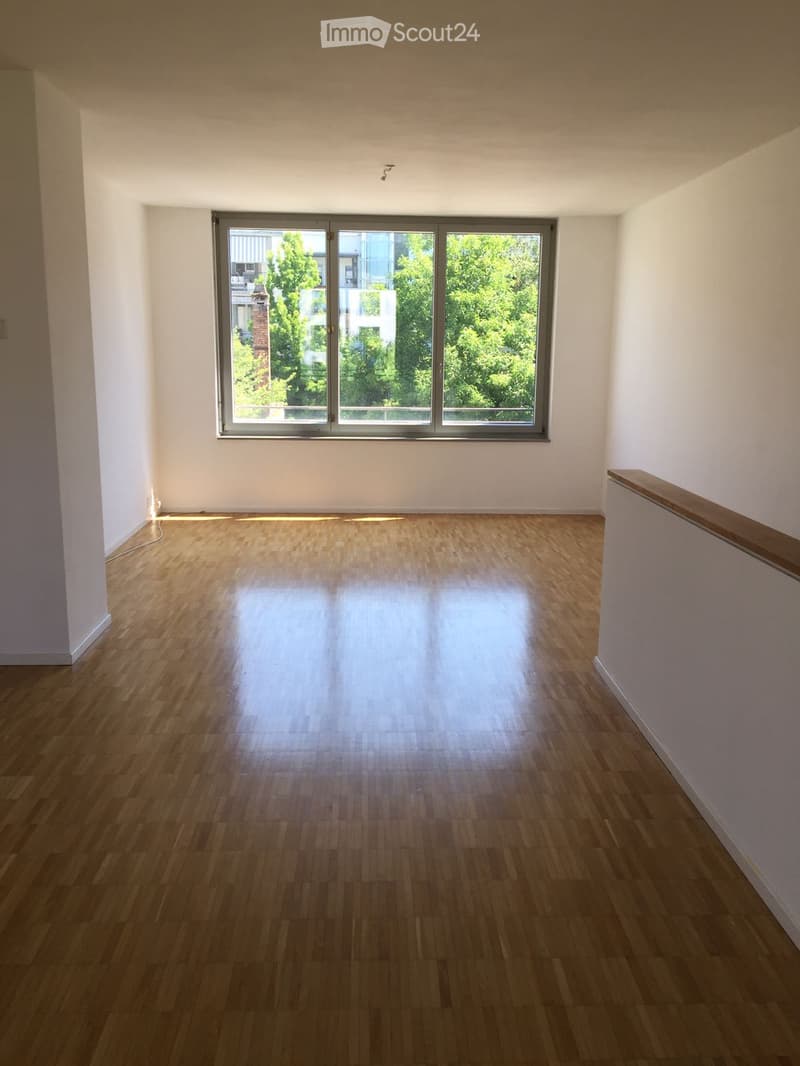Empty room with wooden flooring and large windows, featuring white walls and a small balcony railing visible on the right side.