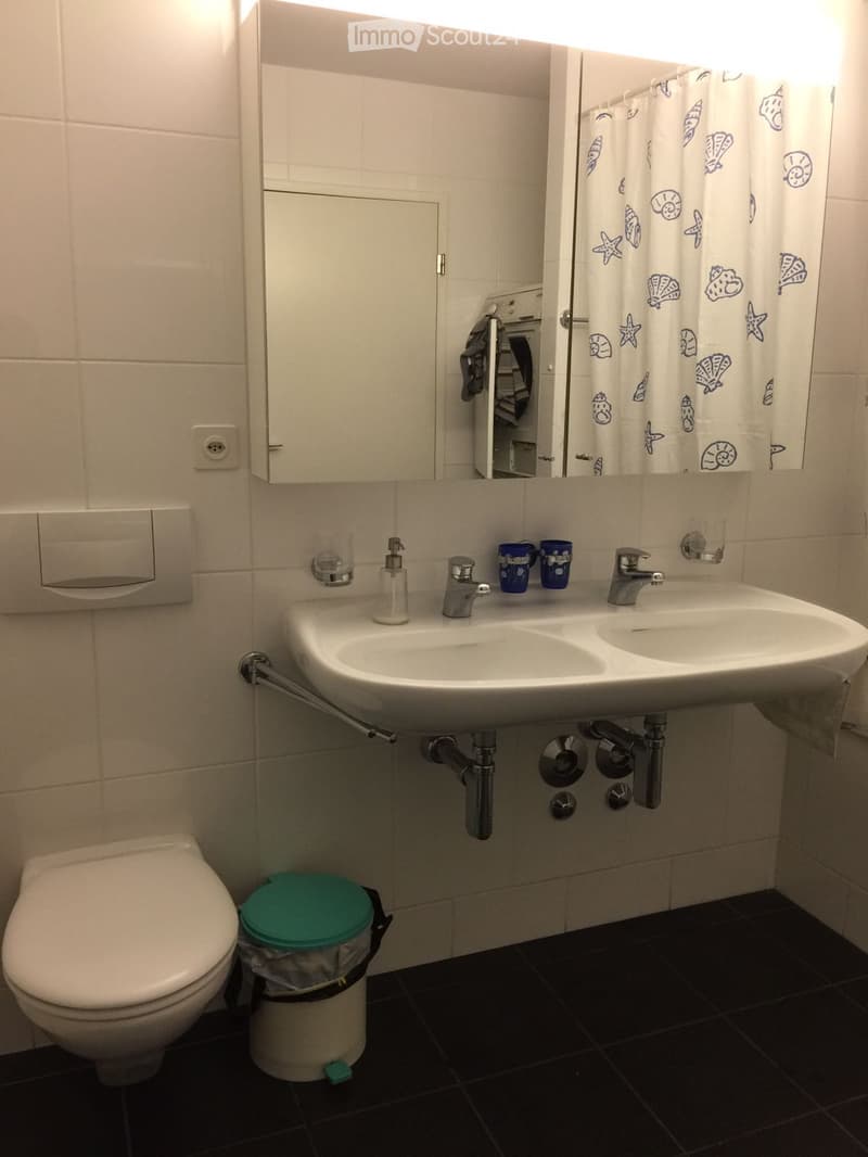 Modern bathroom with a double sink, toilet, mirror, and a small trash bin. The sink has two faucets and two cups placed on it. The walls are tiled, and the floor is dark.
