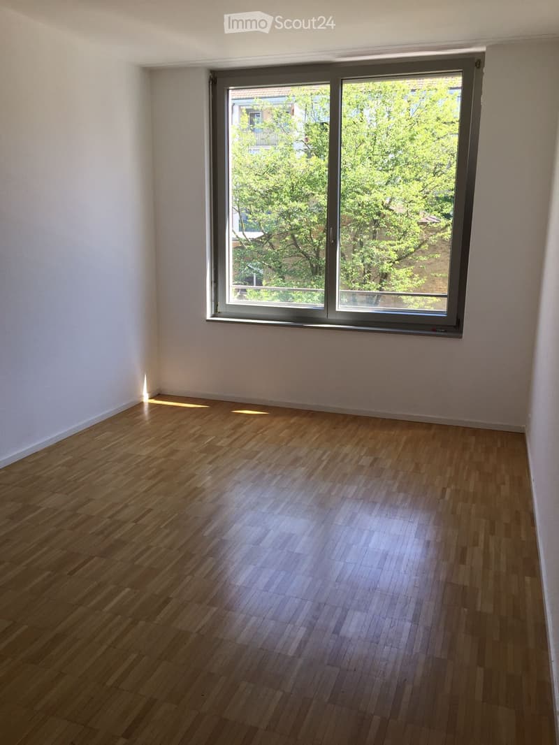 Empty room with wooden flooring and large sliding glass window showing greenery outside.