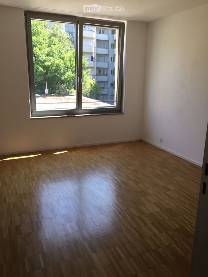 Empty room with wooden flooring and large sliding glass doors leading to a balcony with a view of greenery and neighboring buildings.