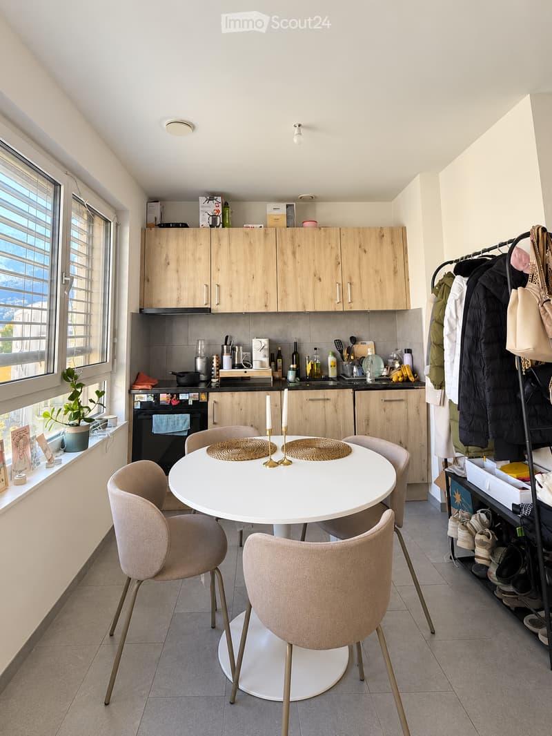 The image shows a modern, well-lit kitchen and dining area. The space features light wooden cabinets, a white dining table with four chairs, and various kitchen appliances and items on the counter. There is also a coat rack with jackets and bags, and a sm