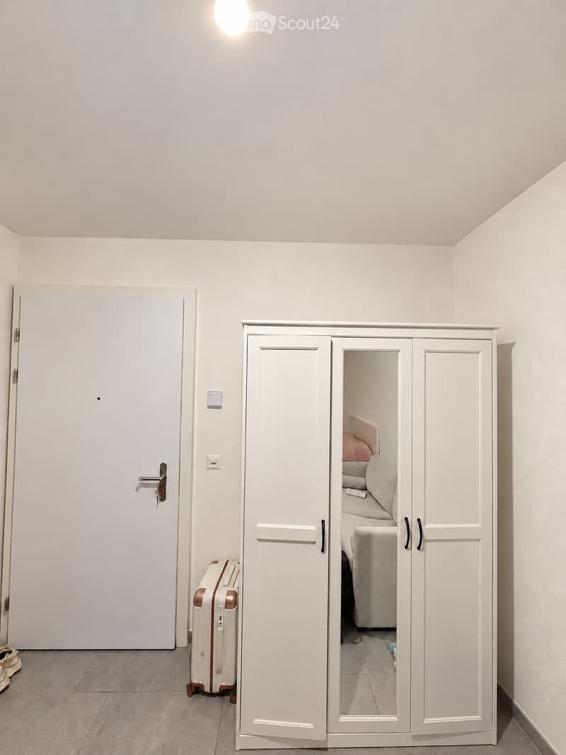 Empty room with white walls, a large white wardrobe with mirrored doors, a closed door, a suitcase, and a pair of shoes on the floor.