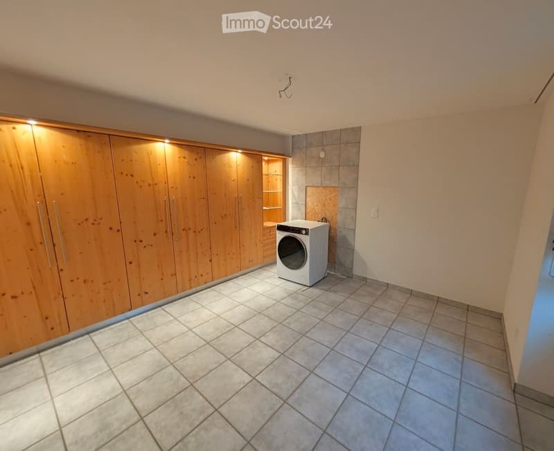 A clean, unfurnished room with tiled flooring, a white washing machine, and a large wooden wardrobe with built-in lighting.