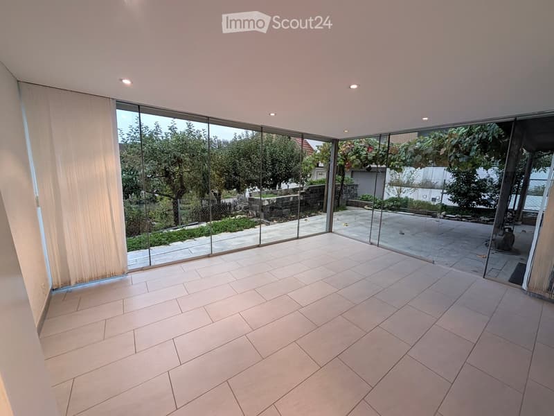 Empty room with large glass sliding doors leading to a garden, tiled floor, and white walls.