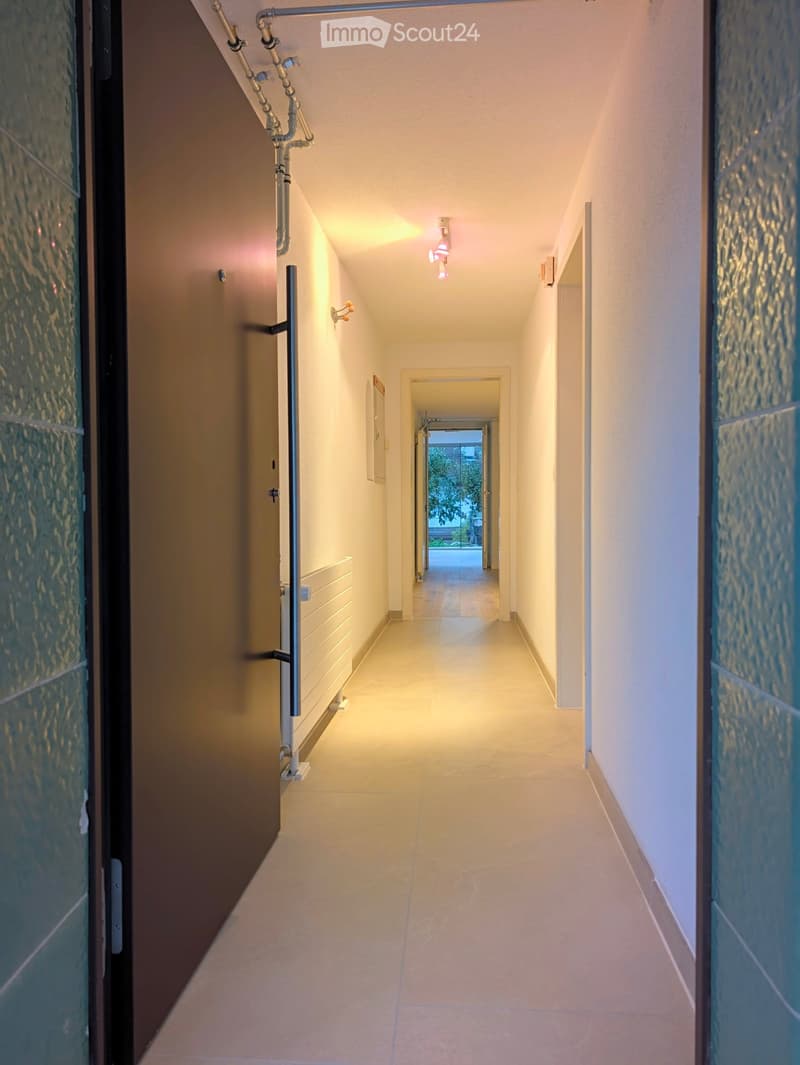 Clean, well-lit hallway with beige tiled flooring, white walls, and modern lighting fixtures. Features a door on the left and a glimpse of an outdoor area at the end.