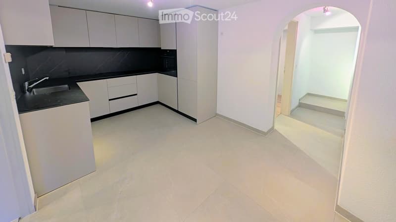 Empty kitchen area with white cabinets, black countertops, and a sink, connected to a bright, open space with tiled flooring.