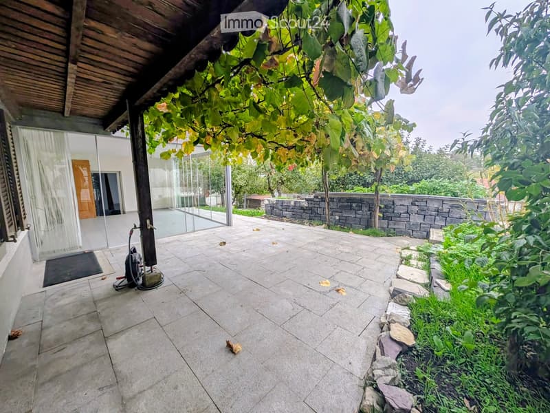 A paved outdoor area with a stone wall, greenery, and a covered entrance to a building.