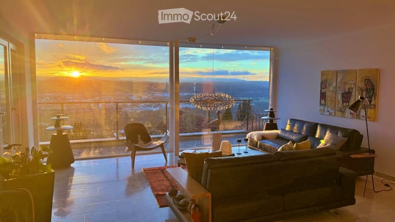 Modern living room with large windows offering a scenic view, featuring a black leather sofa, coffee table, wooden chair, and decorative art pieces.