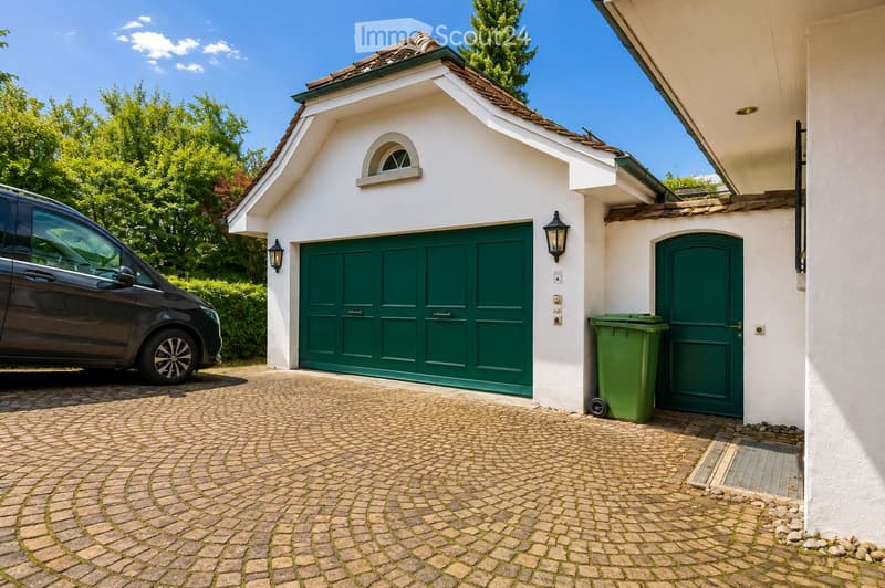 A modern white building with a green garage door, a small arched window above the garage, and a separate green entrance door. A black car is parked nearby, and a green trash bin is placed next to the entrance.