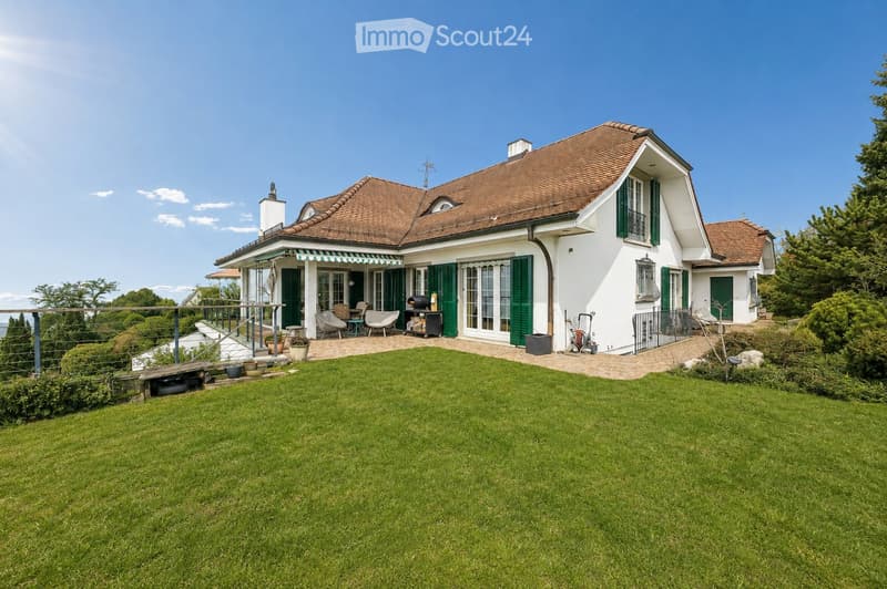 A two-story house with a red-tiled roof, white exterior, and green shutters. The house features a covered porch with outdoor seating, a well-maintained lawn, and a patio area with a grill and outdoor furniture.