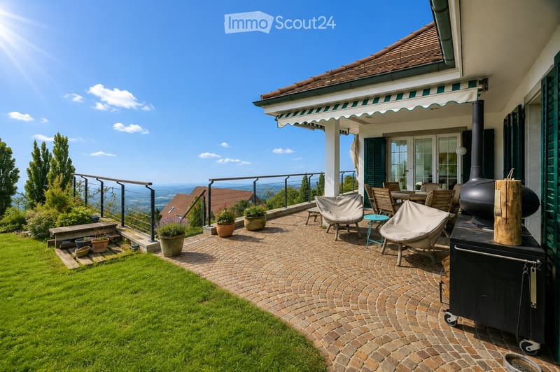 A spacious outdoor patio with a brick-paved floor, featuring lounge chairs, a small table, potted plants, and a grill, overlooking a scenic valley view.