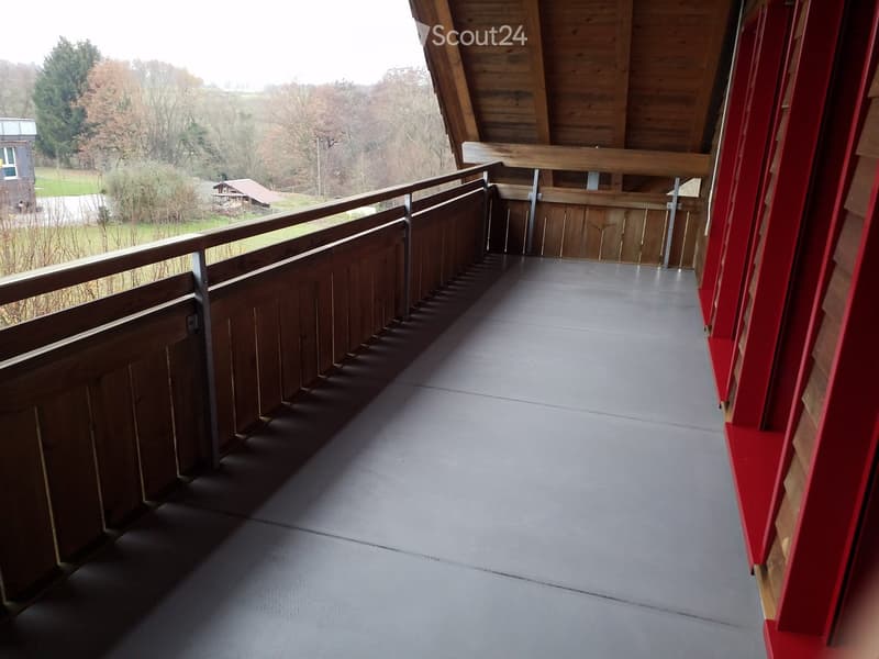 Balcony with wooden railings, grey flooring, and a view of a rural landscape with trees and a small building.