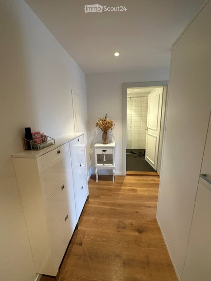 A narrow hallway with light wooden flooring, white walls, and a small white table with a vase of dried flowers. A white cabinet with drawers is on the left side.