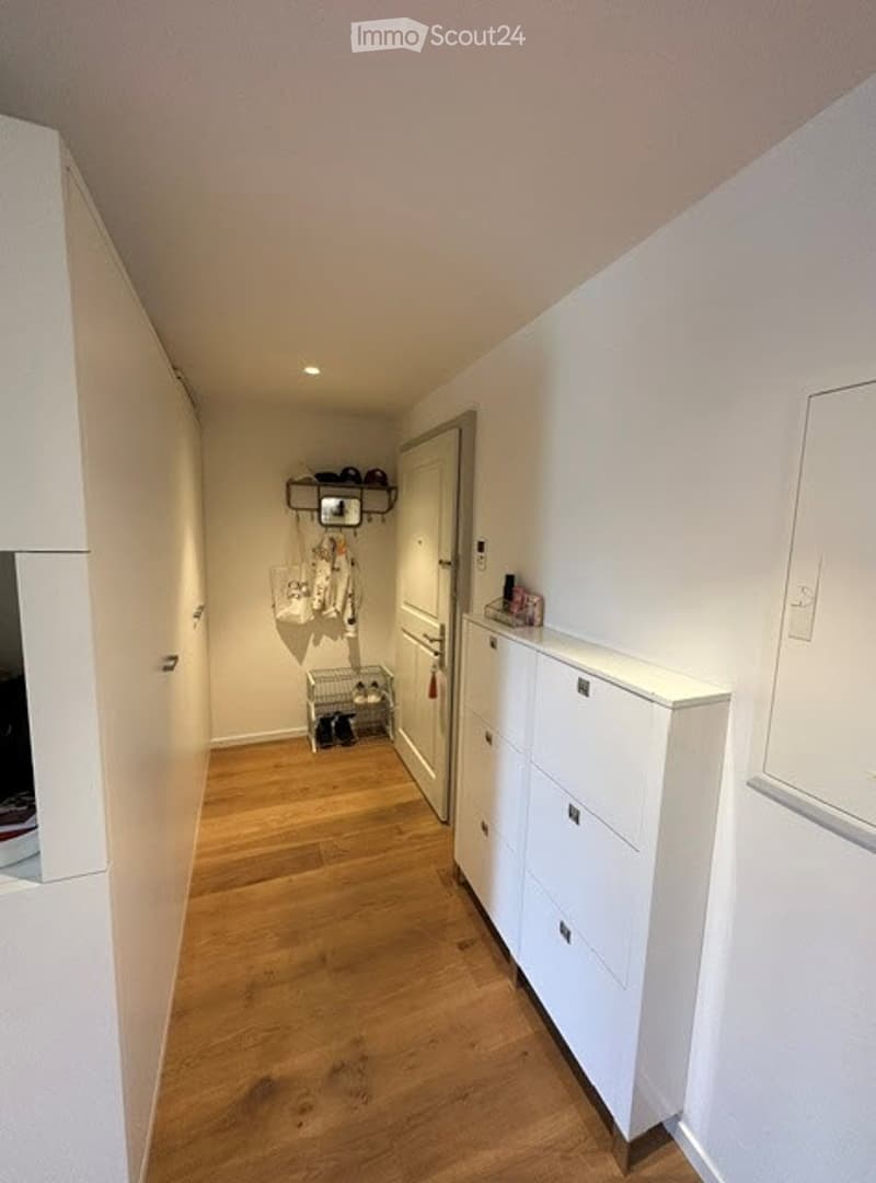 Clean, narrow hallway with light wooden flooring and white walls. Features built-in white storage units on one side and a small shoe rack with coats and bags on the opposite wall.