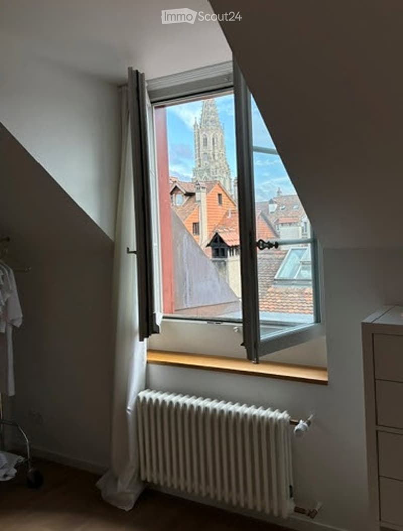 Bright attic room with large window offering a view of a historic building and rooftops. Features wooden floor, radiator, and simple white walls.