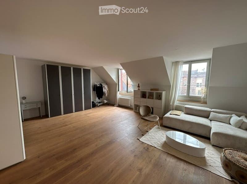 Bright, open living space with wooden flooring, large windows, a grey sectional sofa, a white coffee table, a single chair, a tall wardrobe, and a small shelf unit.