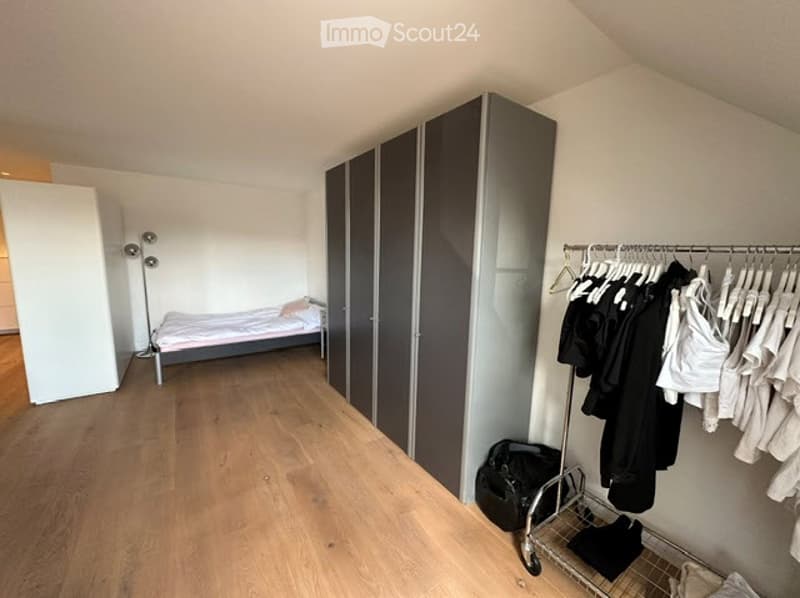 A modern, minimalist bedroom with a single bed, a tall wardrobe, a clothing rack with hanging clothes, a floor lamp, and a backpack on the floor.