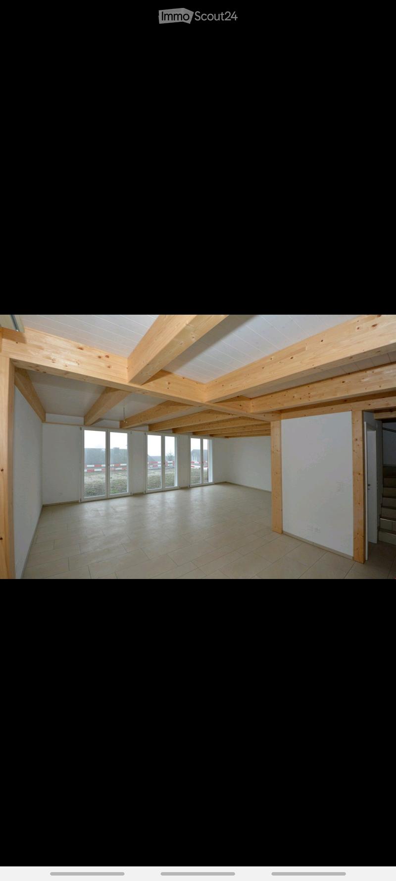 Empty room with exposed wooden beams, large windows, and tiled floor.