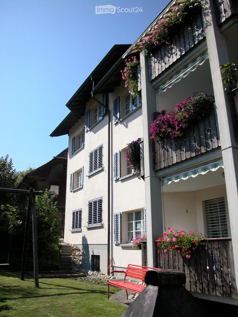 A two-story house with white walls, multiple windows, and flower-filled balconies. A red bench is placed on the grass in front of the house.
