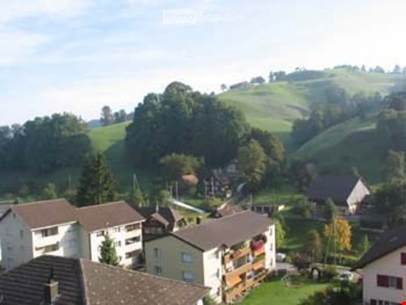 A cluster of residential buildings in a hilly, green landscape with scattered trees and a clear sky.