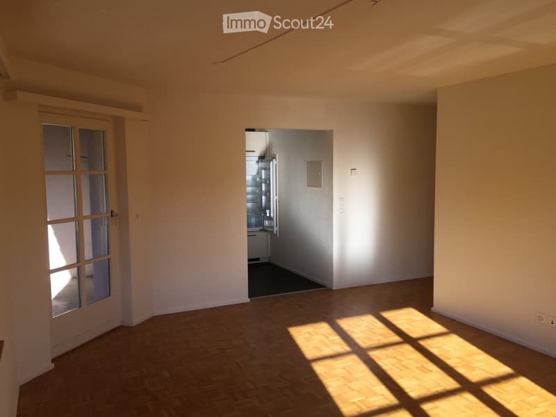 Empty room with wooden flooring and white walls, featuring a glass door and an open kitchen area with a refrigerator visible.