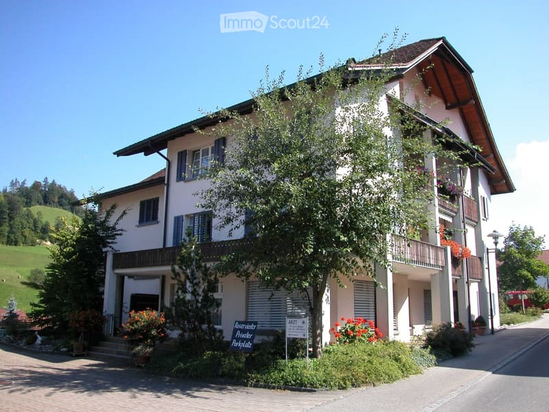 Two-story house with white walls, dark shutters, balconies, and a tree in front. A sign indicates a private parking space.