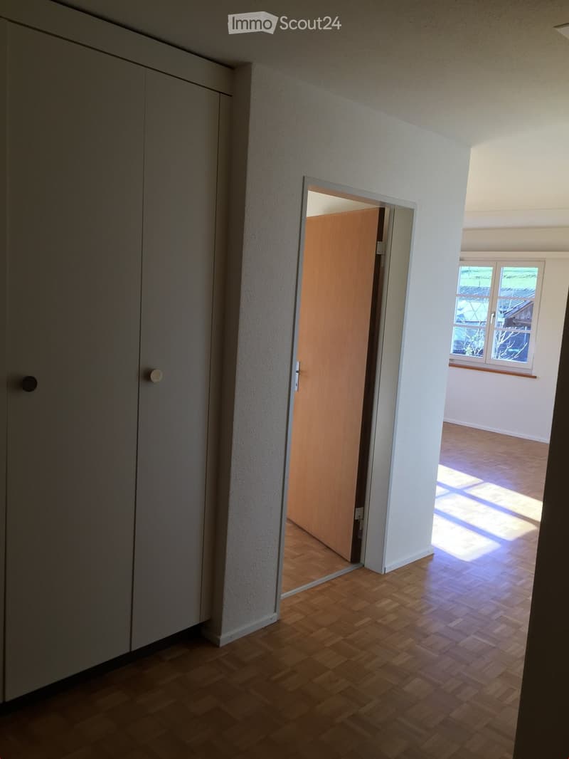 Empty hallway with wooden flooring, white walls, and a partially open wooden door leading to another room with a window showing outdoor view.