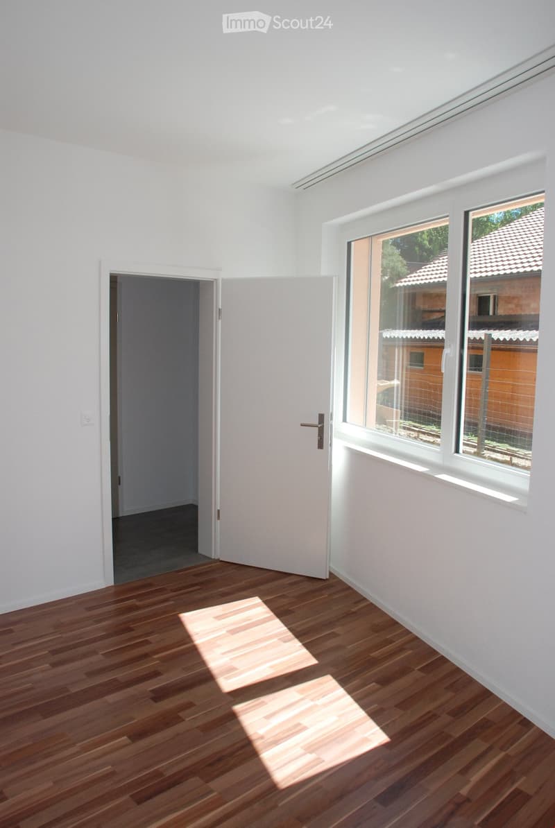 Empty room with wooden flooring, white walls, and large windows showing a view of a garden area. The room has a door leading to another space.