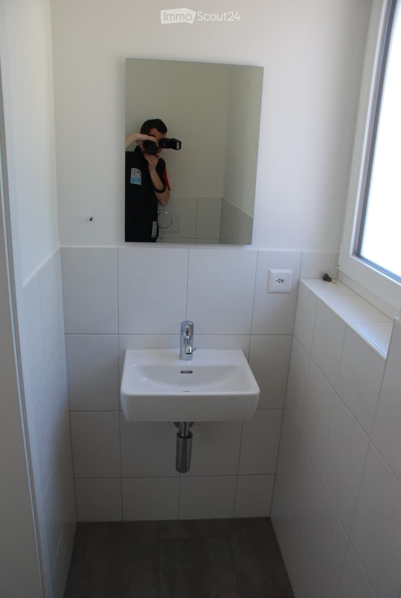 Modern bathroom with a white sink, chrome faucet, and large mirror above it. The walls are tiled in light gray, and the floor is dark gray.