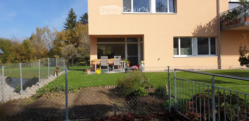 Well-maintained garden with a fenced yard, a patio area with a table and chairs, and a modern building with a balcony in the background.