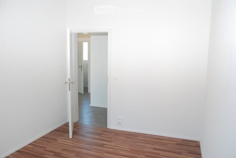Empty room with wooden flooring and white walls, featuring an open doorway leading to another room with tiled flooring.