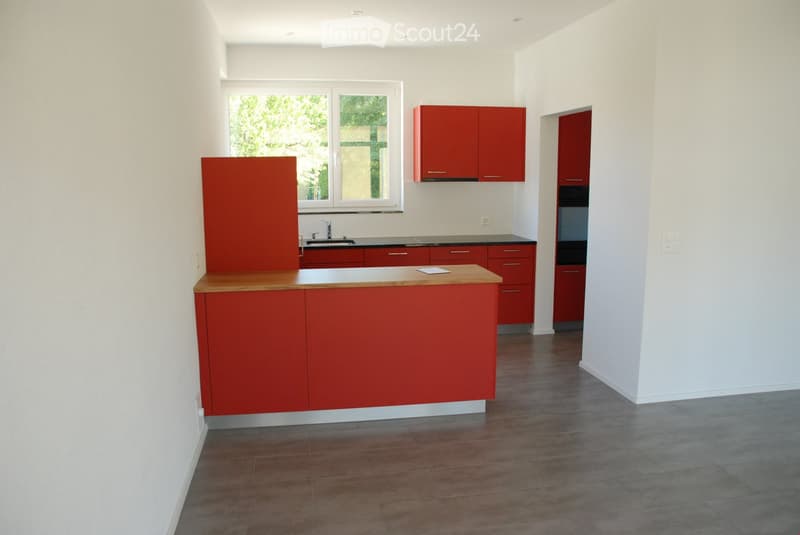 Modern kitchen with red cabinets, a central island, and a sink area. The space features a large window with a view of greenery outside.