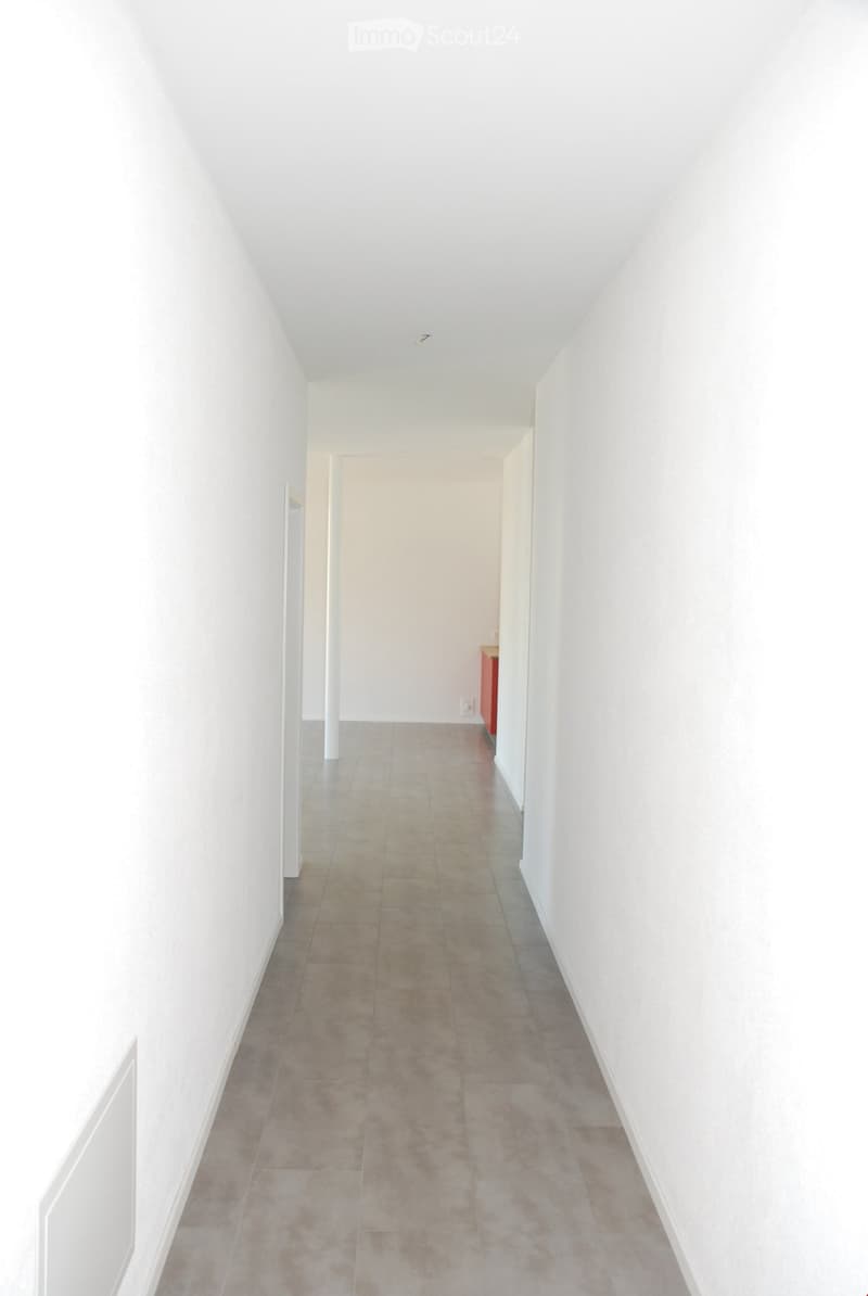 A long, narrow hallway with white walls and beige tiled flooring, leading to an open doorway at the end.