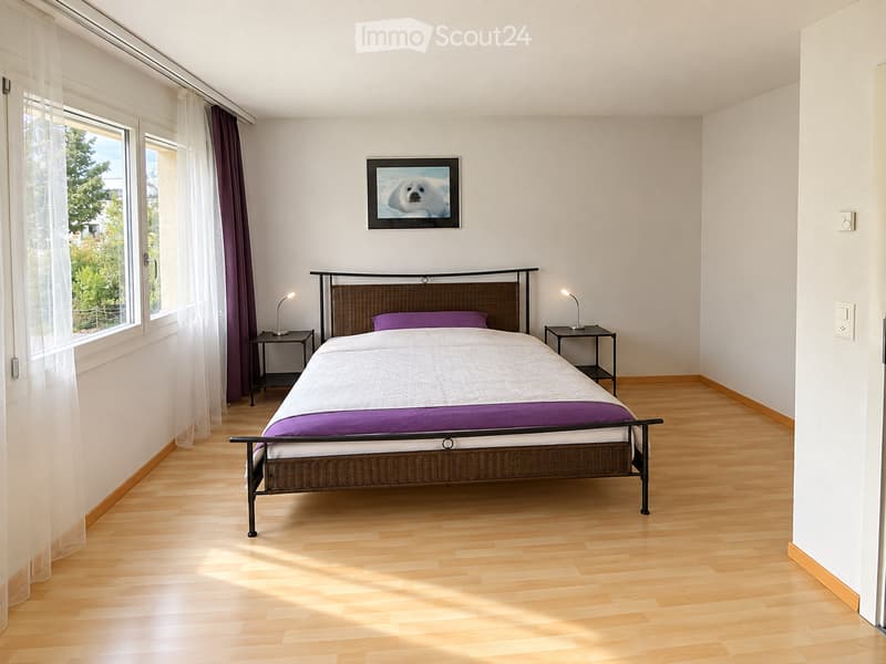 Bright bedroom with wooden flooring, a double bed with white and purple bedding, two nightstands with lamps, and a picture frame on the wall.
