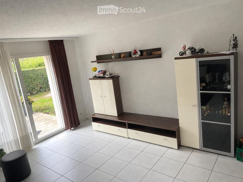 A modern living room with white tiled flooring, large window with brown curtains, a grey stool, a white and brown cabinet, a wooden shelf with decorative items, and a tall cabinet with glassware and bottles.