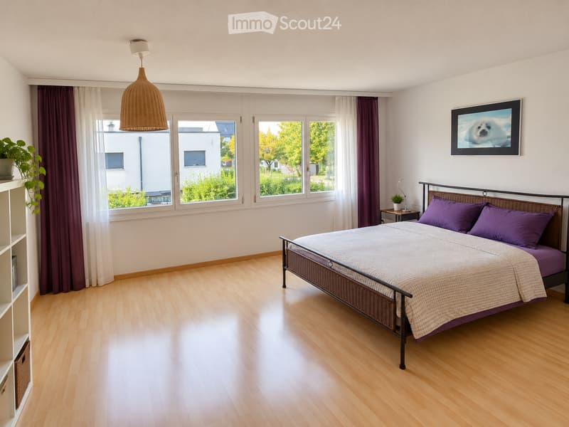 Bright bedroom with wooden flooring, large windows, a bed with purple bedding, a nightstand with a lamp, a potted plant, and a shelf with books.