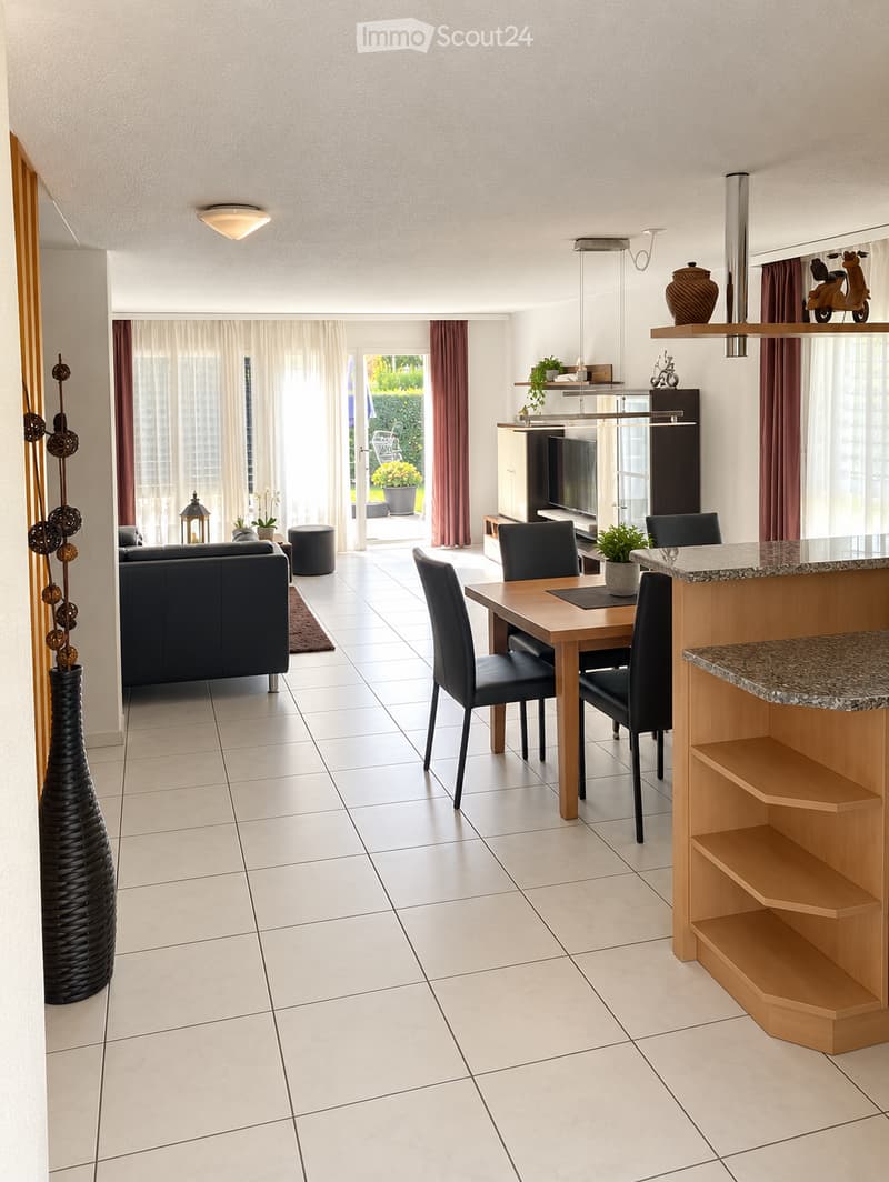 Open-plan living and dining area with a sofa, dining table, chairs, a stool, a vase, a television, shelves, and plants. The space features large windows with curtains and a view of a garden.