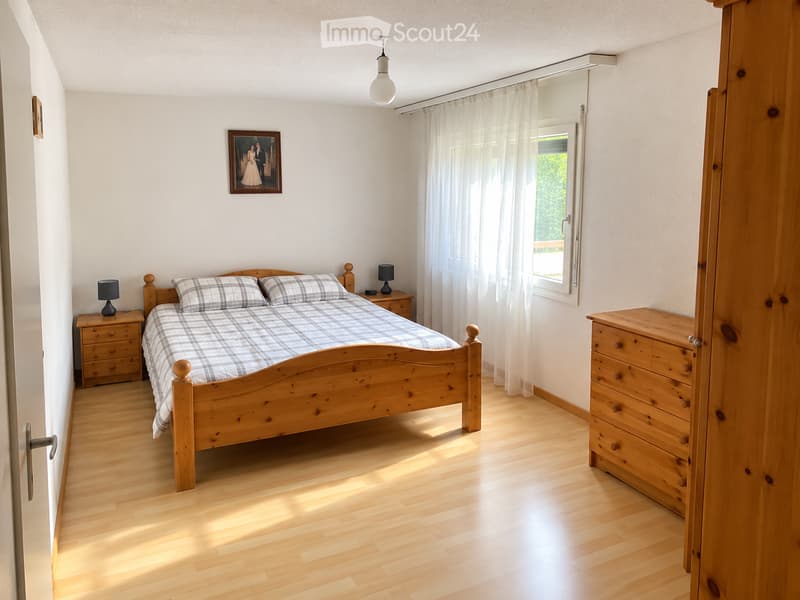 Cozy bedroom with wooden bed, plaid bedding, nightstands, lamps, and a dresser. Natural light from a window with white curtains.