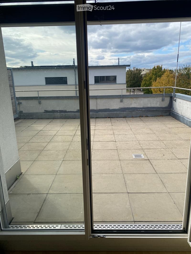 Empty balcony with tiled floor, metal railing, and a view of surrounding buildings and trees.