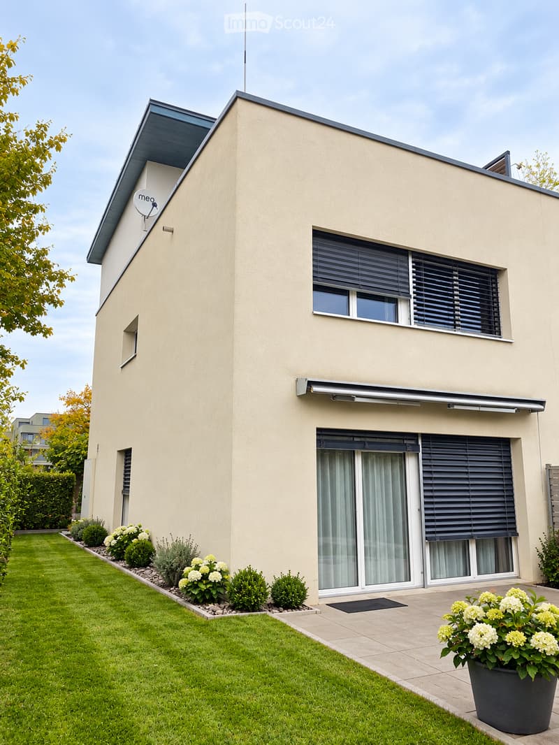 Modern two-story house with beige exterior, large windows, sliding glass doors, and a well-maintained garden with shrubs and flowers.