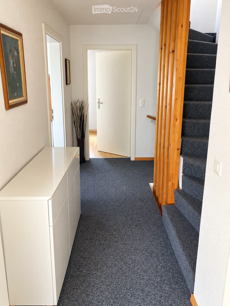 Clean hallway with carpeted floor, white walls, a staircase with wooden railing, a white console table, and a vase with dried plants.